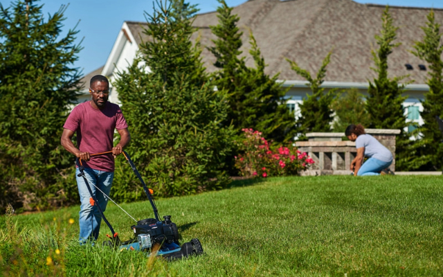 Man walking behind mower on grassy slope as woman gardens in the background.