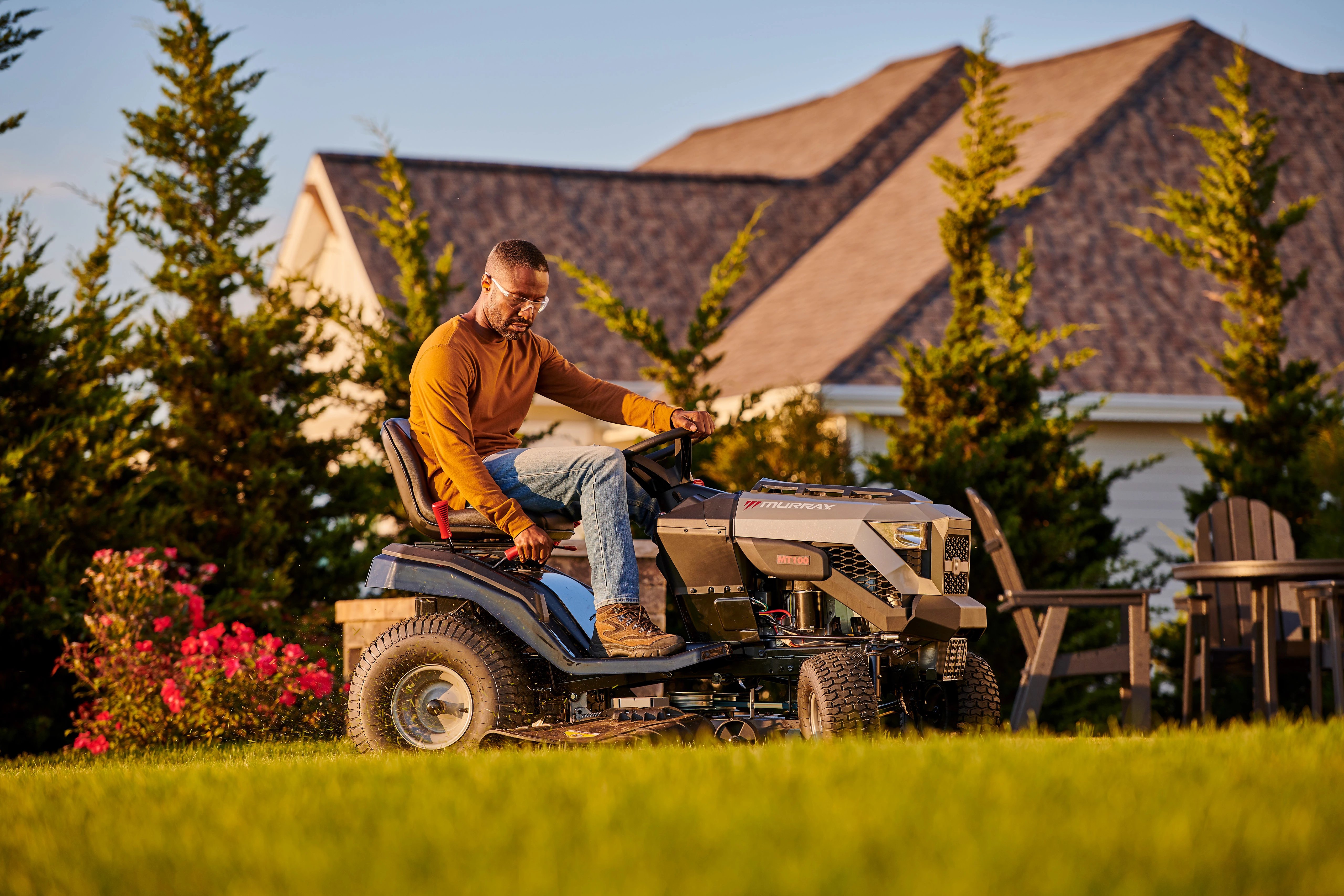 A man wearing safety glasses driving a Murray MT100 riding lawn mower across a sunny residential lawn, powered by a Briggs & Stratton engine.